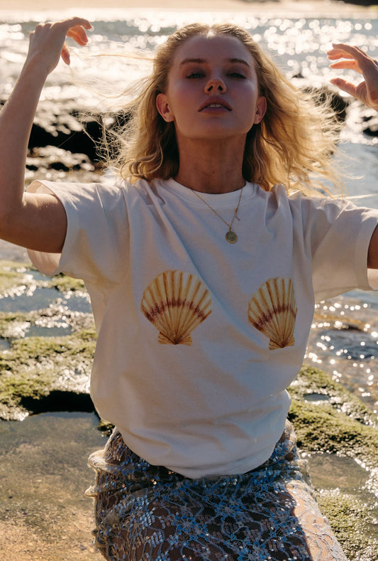 Woman wearing a white t-shirt with shell designs by a body of water.