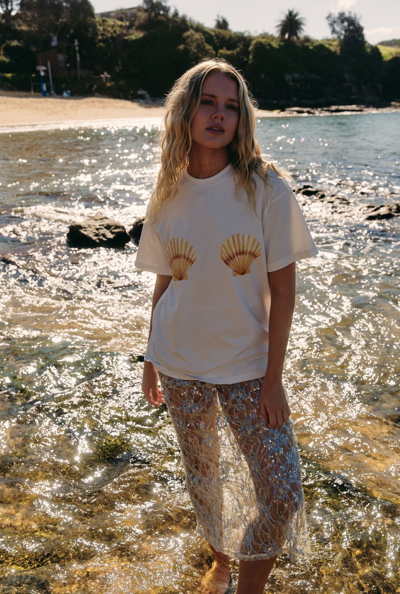 Woman standing by a body of water wearing a white t-shirt with shell design.